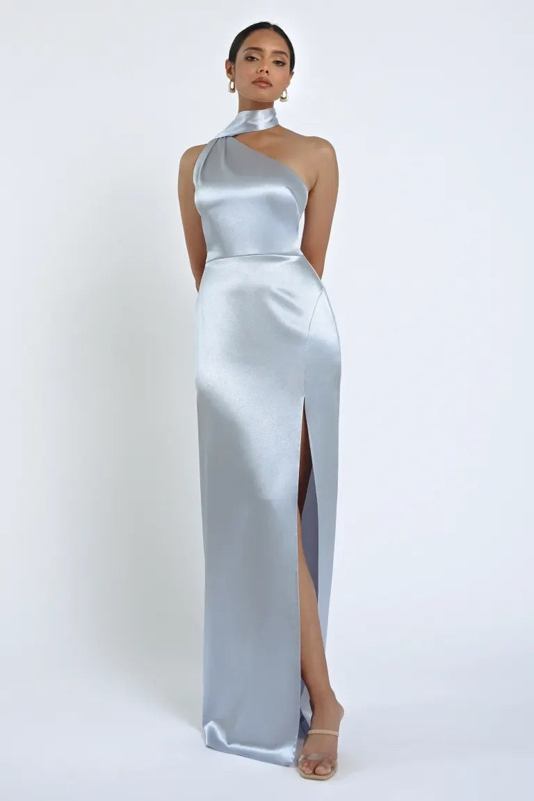 A woman stands wearing the Hepburn Bridesmaid Dress by Jenny Yoo, a sleeveless, silver satin back crepe evening gown with a high slit from Bergamot Bridal, exuding Hollywood glam. She is posed in front of a plain white background.