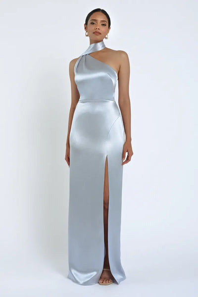 A woman is standing against a plain backdrop wearing the Hepburn Bridesmaid Dress by Jenny Yoo, a silver satin back crepe, one-shoulder gown with a high slit from Bergamot Bridal that exudes Hollywood glam.