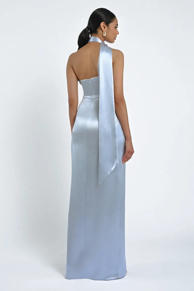 A woman is wearing the Hepburn Bridesmaid Dress by Jenny Yoo from Bergamot Bridal, a long, light blue satin gown with a halter neck and a long ribbon detail at the back. Evoking Hollywood glam, she is seen from behind against a plain white background.