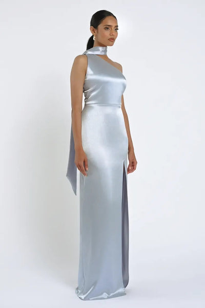 A woman stands wearing the Hepburn Bridesmaid Dress by Jenny Yoo, a sleeveless, shimmering silver gown featuring a high collar and a thigh-high slit, exuding Hollywood glam against a plain white background. This exquisite piece is from Bergamot Bridal.