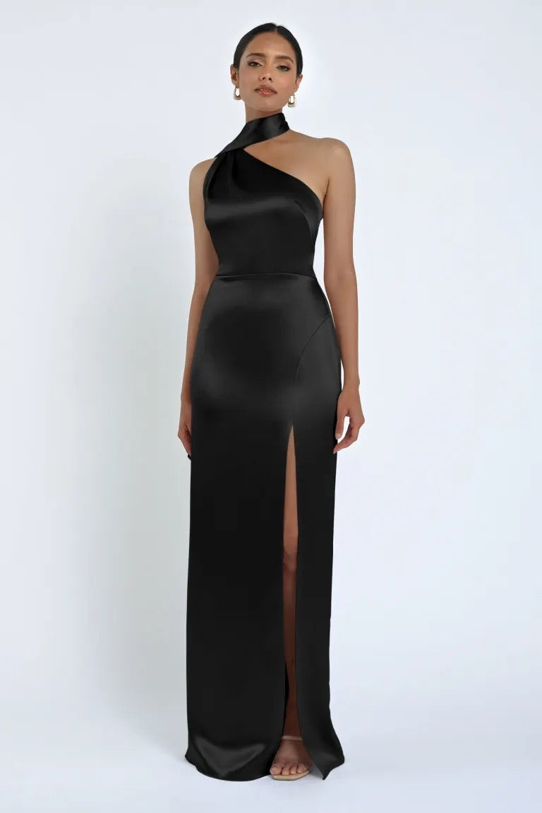 A woman stands wearing the Hepburn Bridesmaid Dress by Jenny Yoo from Bergamot Bridal, a sleeveless, black satin back crepe gown with a high slit and a halter neckline, exuding classic Hollywood glam.