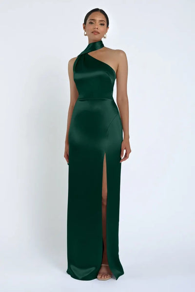 A woman stands in the Hepburn Bridesmaid Dress by Jenny Yoo from Bergamot Bridal, a stunning full-length emerald green satin back crepe dress with a high slit and halter neck, exuding Hollywood glam against a plain background.