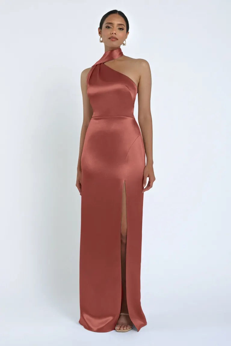 A person is wearing a floor-length, rust-colored Hepburn Bridesmaid Dress by Jenny Yoo from Bergamot Bridal, featuring a high neckline and a thigh-high slit. Posing against a plain white background, the satin back crepe fabric enhances the gown with an extra touch of Hollywood glam.