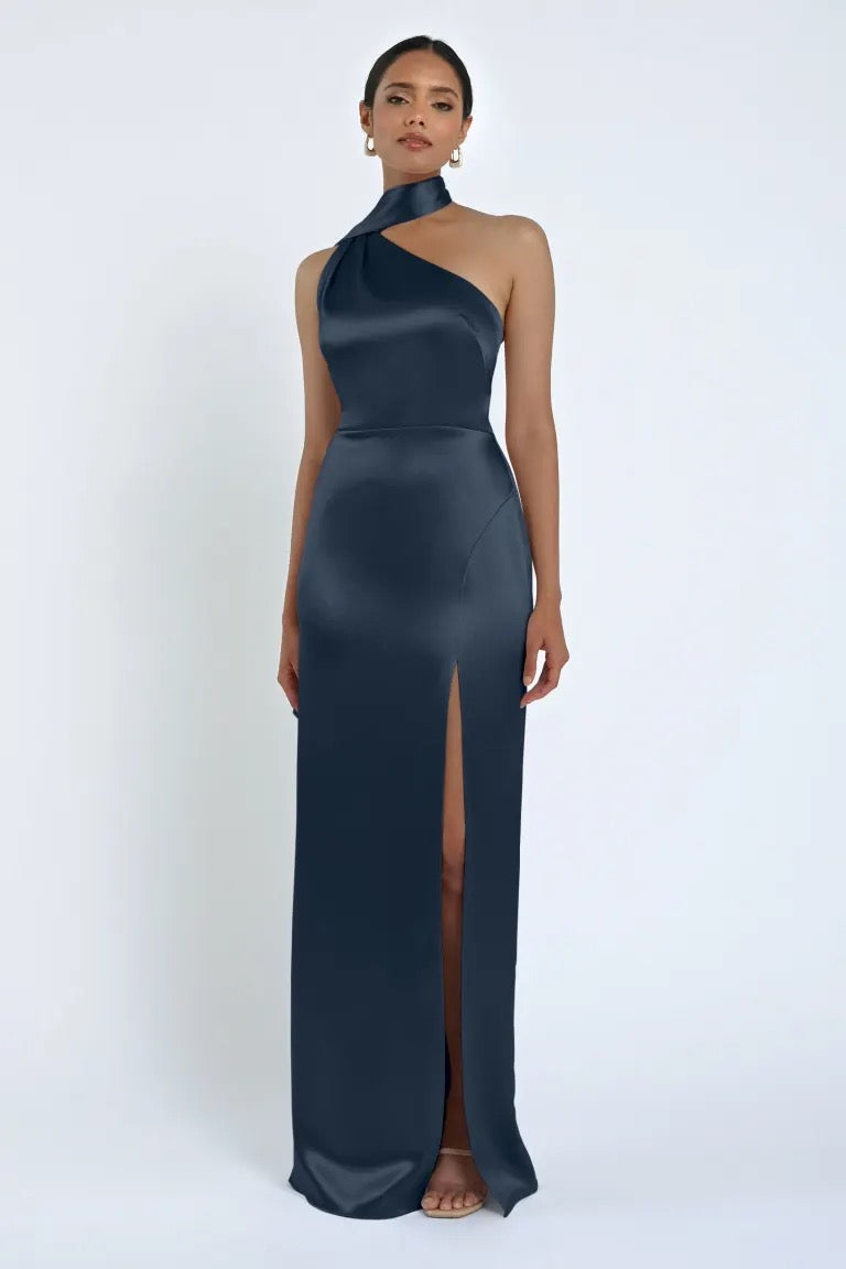 A woman is standing in the Hepburn Bridesmaid Dress by Jenny Yoo from Bergamot Bridal, a sleeveless, dark teal satin back crepe evening gown with a high slit on one side. The floor-length dress features a high neckline reminiscent of classic Hepburn elegance, and she is wearing earrings.