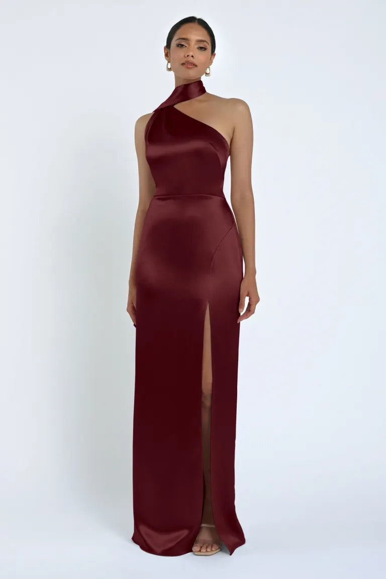 A woman stands wearing the sleeveless Hepburn Bridesmaid Dress by Jenny Yoo for Bergamot Bridal—a burgundy satin gown with a high slit and halter neckline—against a plain background, exuding Hollywood glam.