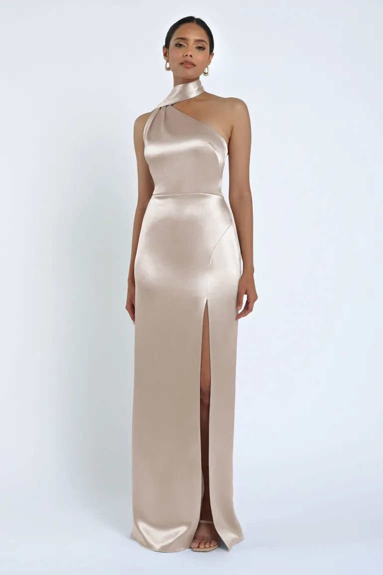 A person stands wearing the Hepburn Bridesmaid Dress by Jenny Yoo, a floor-length, Hollywood glam one-shoulder dress with a high slit, crafted from luxurious satin back crepe by Bergamot Bridal.