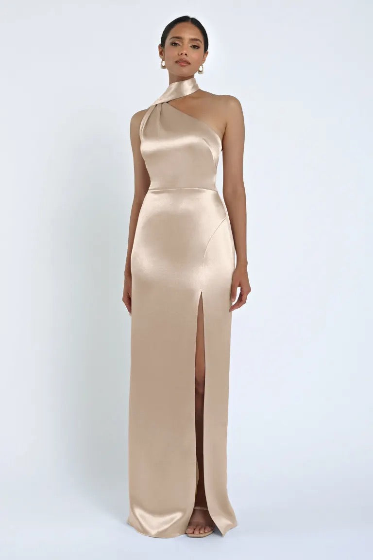 A person wearing the Hepburn Bridesmaid Dress by Jenny Yoo from Bergamot Bridal, a long gold satin back crepe one-shoulder dress with a high slit, stands against a plain white background, exuding Hollywood glam.