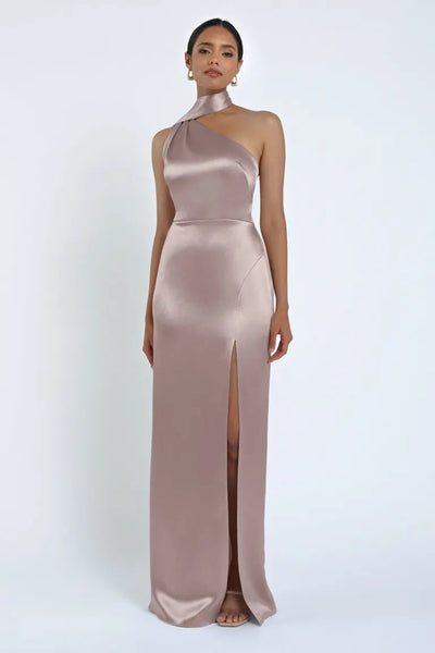 A person in the Hepburn Bridesmaid Dress by Jenny Yoo from Bergamot Bridal, a taupe sleeveless satin gown with a high slit that evokes classic Hollywood glam, stands elegantly against a plain white background.