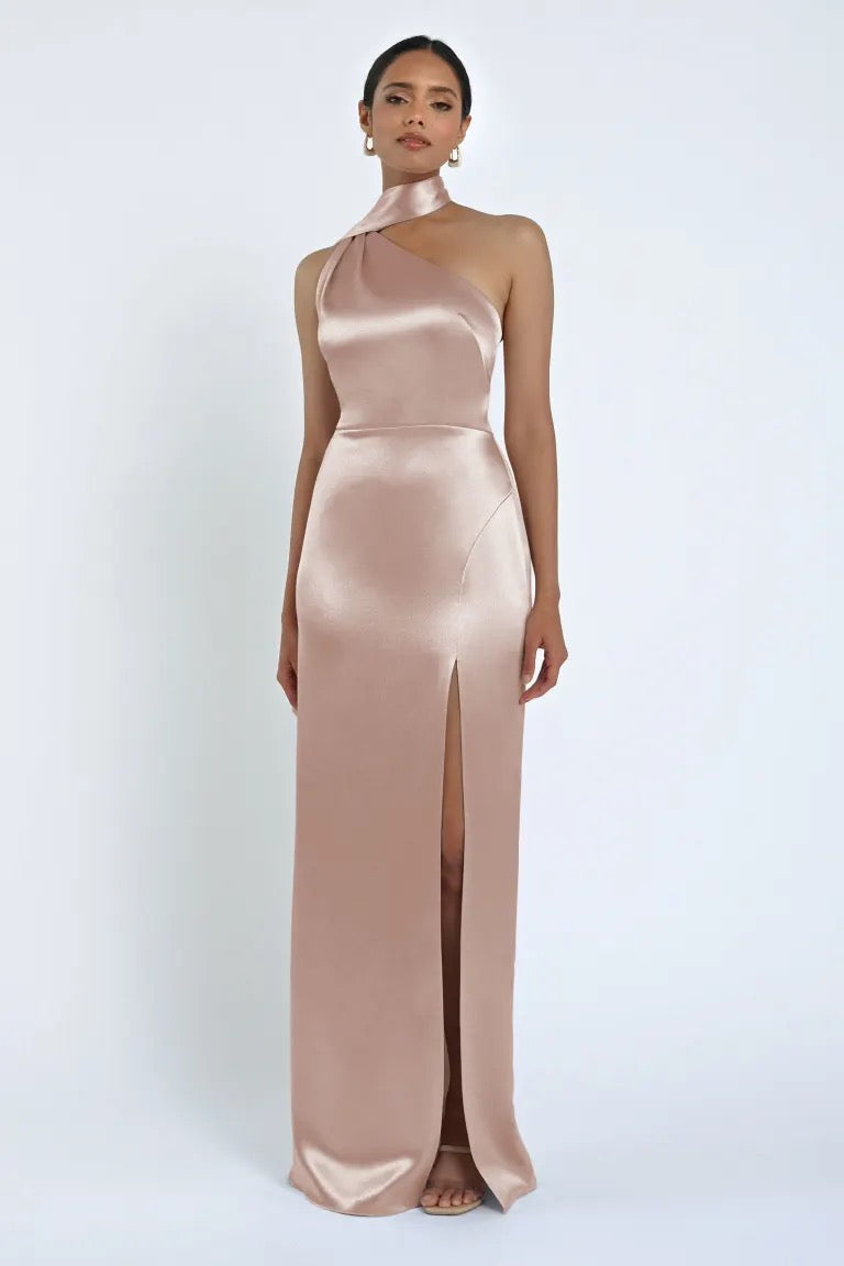 A woman is wearing the Hepburn Bridesmaid Dress by Jenny Yoo from Bergamot Bridal, a floor-length, one-shoulder satin back crepe gown in a light pink hue with a thigh-high slit, exuding Hollywood glam.