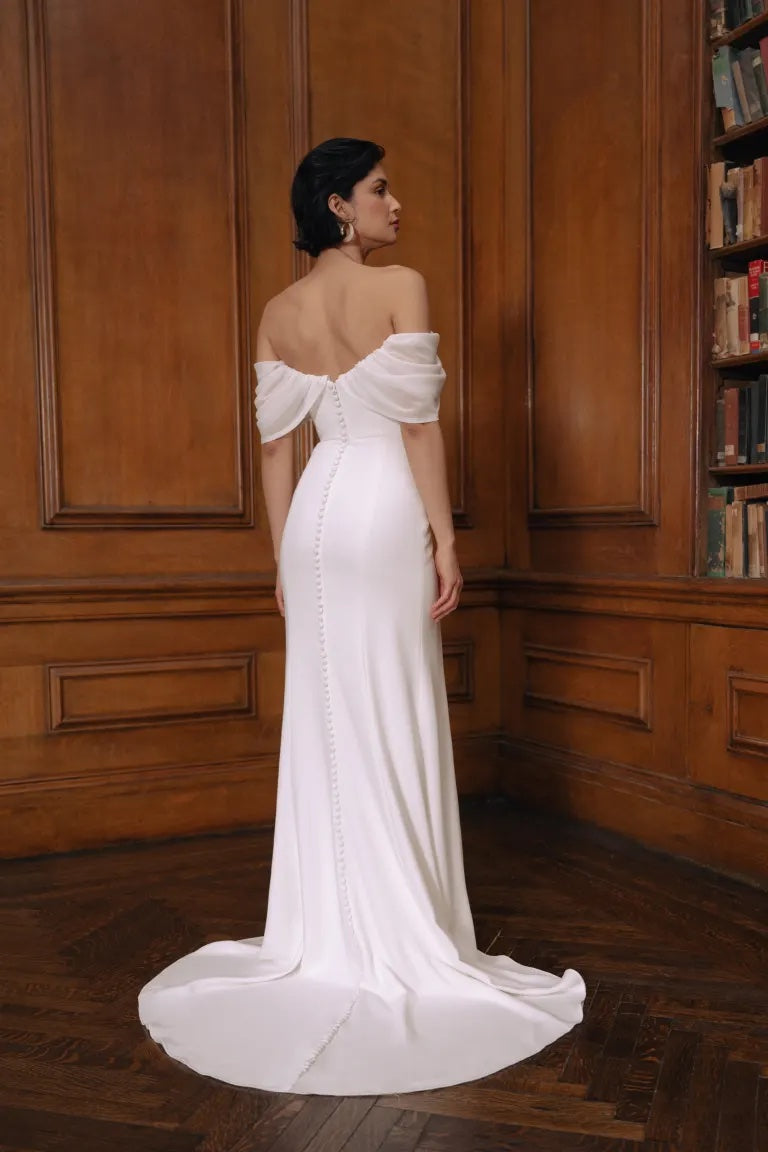 A woman in an elegant Jenny Yoo Wedding Dress off-the-shoulder gown featuring a sweetheart neckline, standing in a library setting.