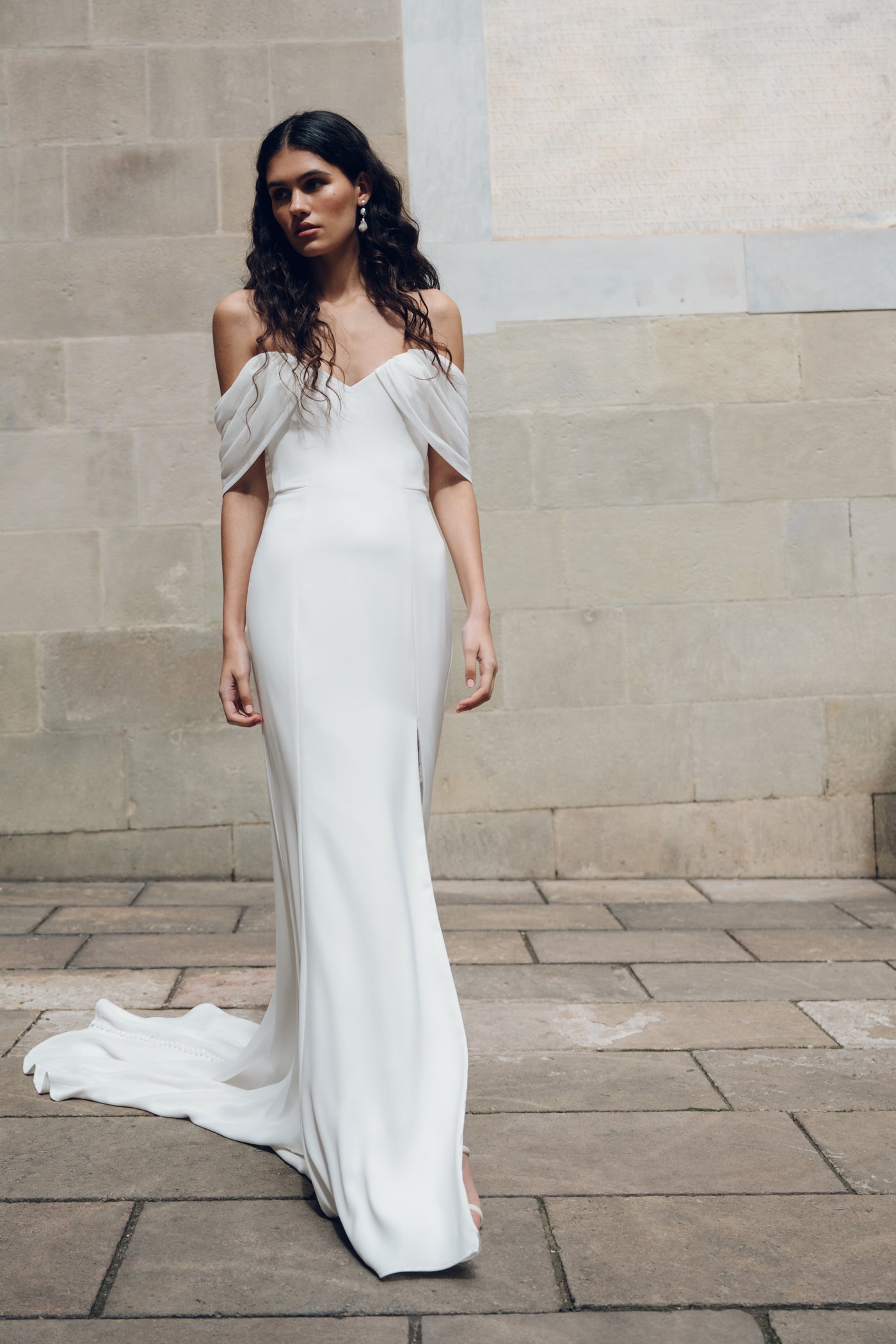 A woman in the Hilaire - Jenny Yoo Wedding Dress by Bergamot Bridal, featuring an off-shoulder design with a fit and flare silhouette, stands on a stone pavement. Her neutral expression enhances the dress's elegance against a timeless stone wall backdrop.