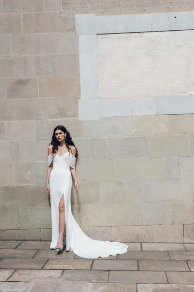 A woman wearing the Hilaire - Jenny Yoo Wedding Dress by Bergamot Bridal, featuring a white fit and flare silhouette with chiffon sleeves and a thigh-high slit, stands against a stone wall.