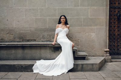 A woman wearing the Hilaire - Jenny Yoo Wedding Dress by Bergamot Bridal, featuring a white off-the-shoulder design with a sweetheart neckline and fit and flare silhouette, sits on a stone bench against a textured wall.