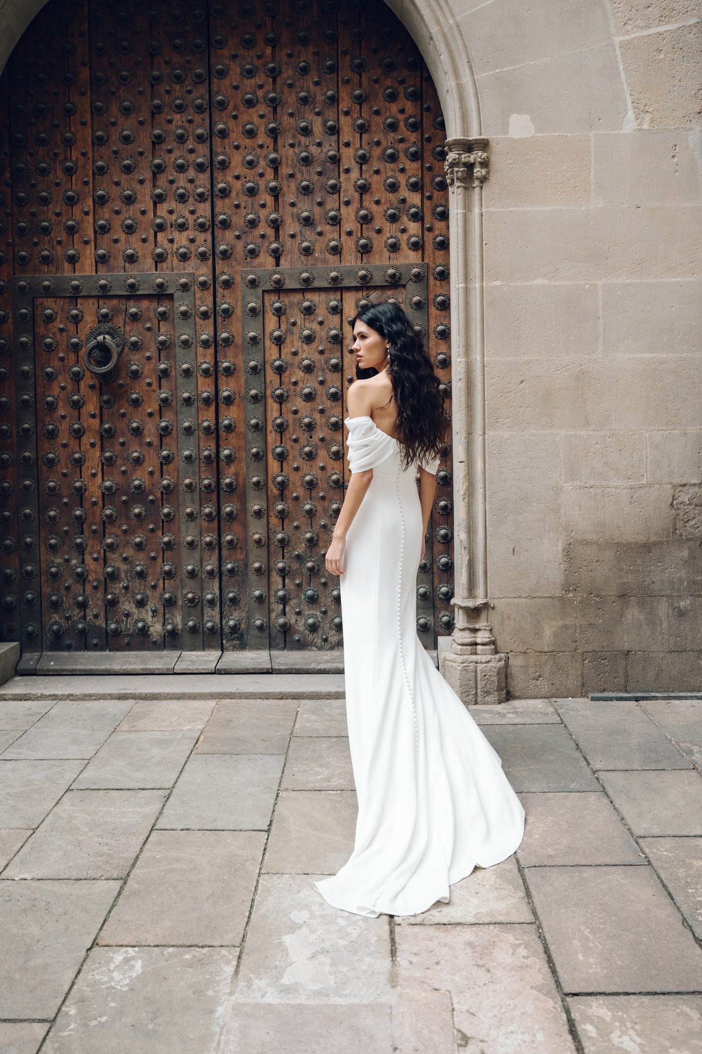 A woman in the Hilaire - Jenny Yoo Wedding Dress by Bergamot Bridal stands gracefully in front of a large, ornate wooden door set in a stone wall. The elegant white dress boasts chiffon sleeves and a fit and flare silhouette, perfectly complementing her poised stance as she faces sideways with her hair gently cascading down.