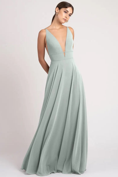 A woman in a flowing sage green gown with a plunging V-neck stands elegantly against a light background, wearing the Hollis Bridesmaid Dress by Jenny Yoo from Bergamot Bridal.