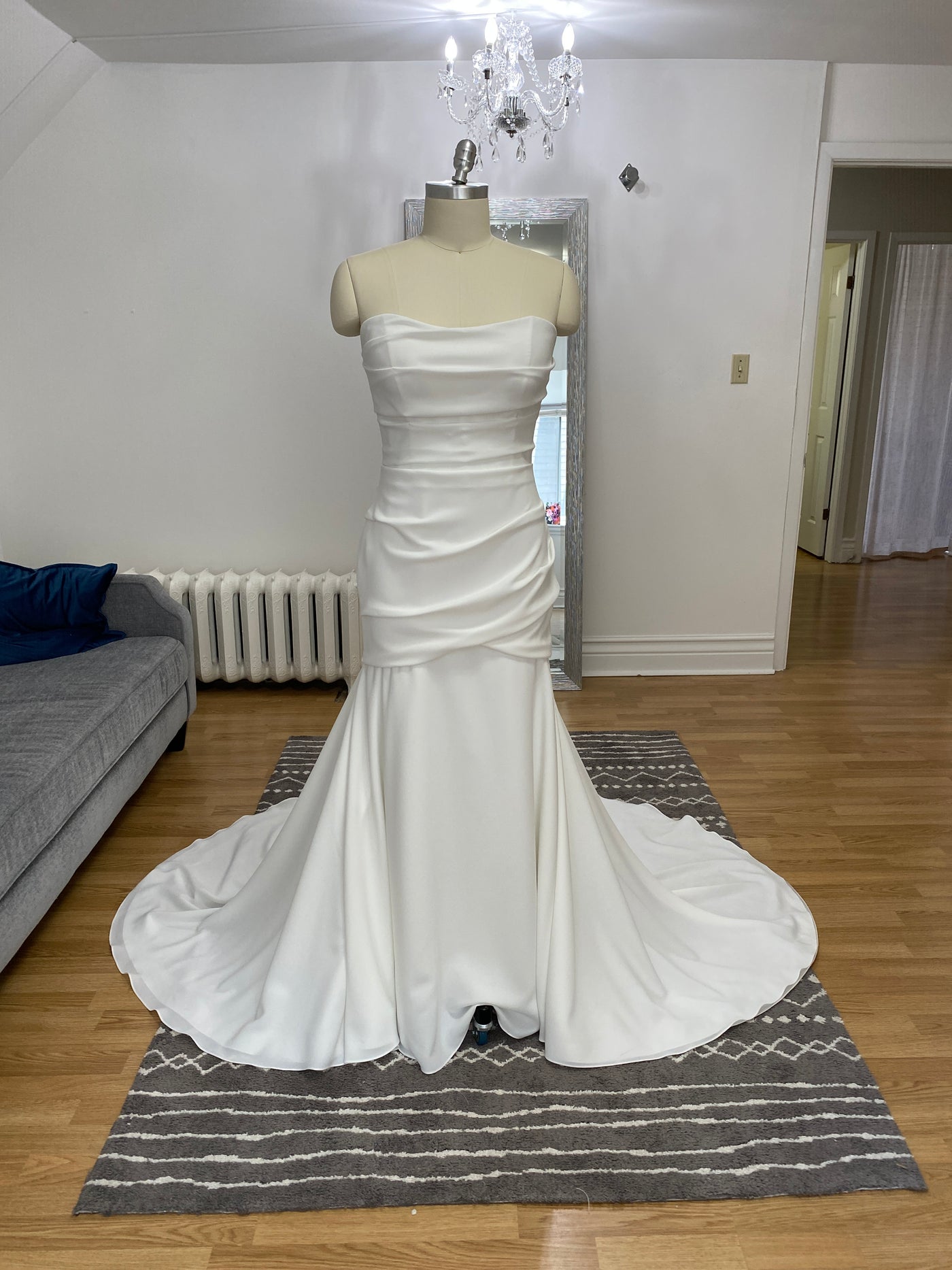 A strapless, white Paloma Blanca Simple Crepe Mermaid Gown Style 4975 - Off The Rack displayed on a mannequin in a room with a gray sofa, patterned rug, and a chandelier at Bergamot Bridal.