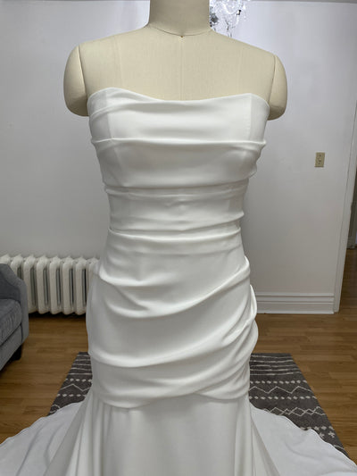 A mannequin displaying a Paloma Blanca Simple Crepe Mermaid Gown Style 4975 - Off The Rack with a draped tunic bodice and flowing skirt in an indoor setting at Bergamot Bridal.