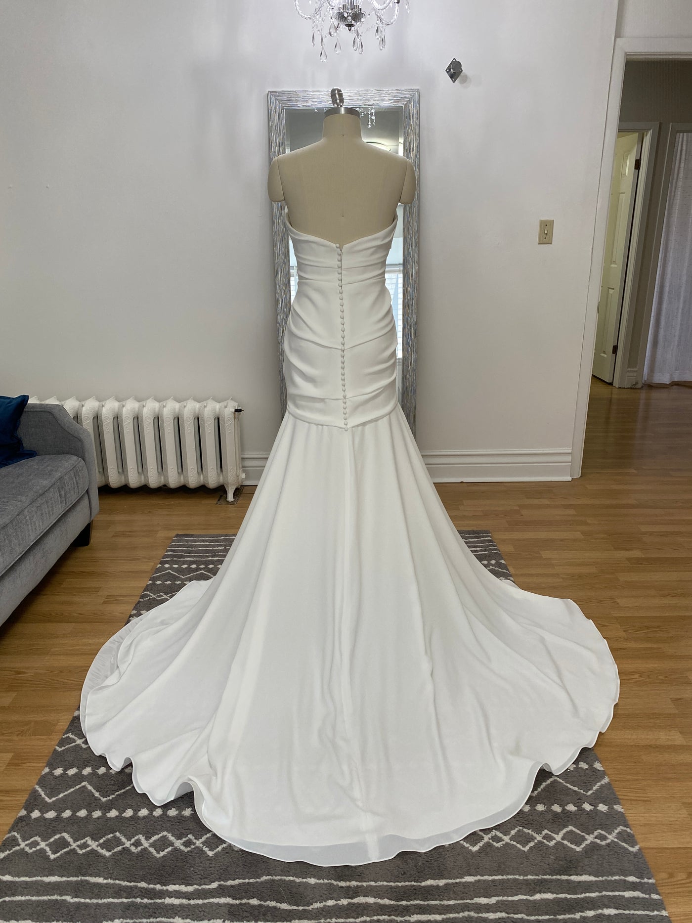 A white Paloma Blanca Simple Crepe Mermaid Gown Style 4975 - Off The Rack wedding dress on a mannequin in a living room with a grey sofa and patterned rug.