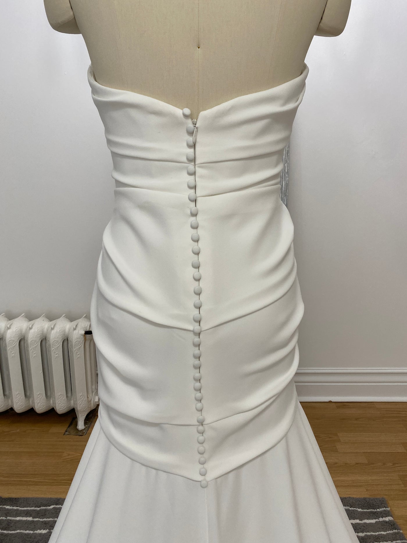 Detail of a white Paloma Blanca Simple Crêpe wedding dress on a mannequin, featuring a ruched waist and a line of decorative buttons down the back from Bergamot Bridal.
