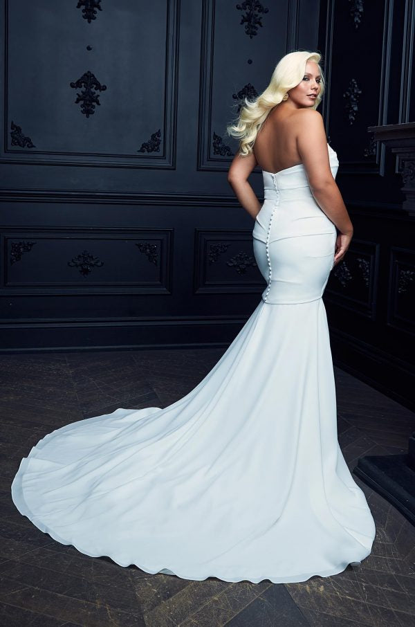 A woman in a Bergamot Bridal Paloma Blanca Simple Crepe Mermaid Gown Style 4975 - Off The Rack with a flowing train, standing in a dark paneled room, looking over her shoulder.