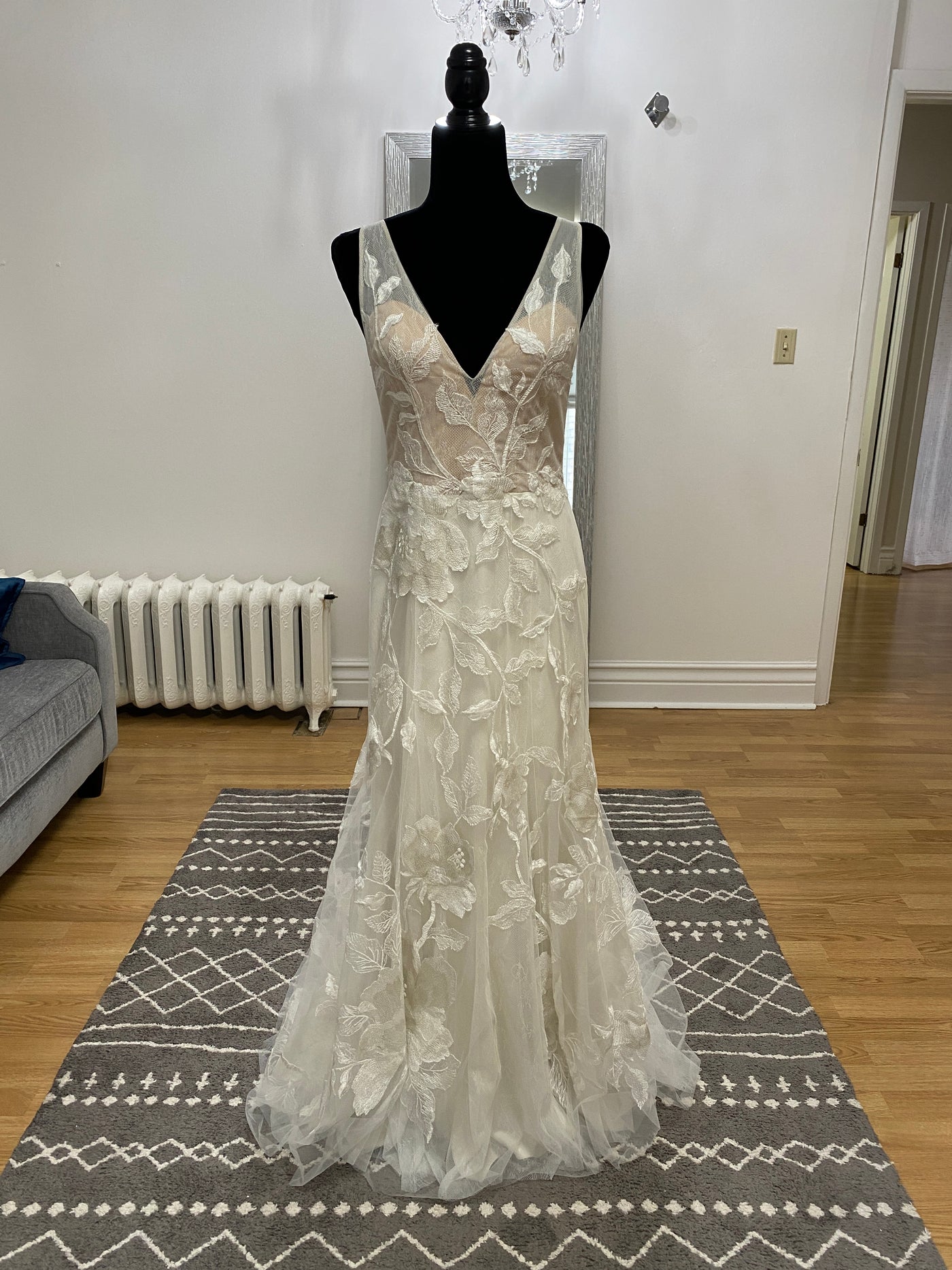 A Willowby by Watter Honor 52122 - Sample Size white floral lace wedding dress on a mannequin in a room with a geometric rug and light wooden flooring.