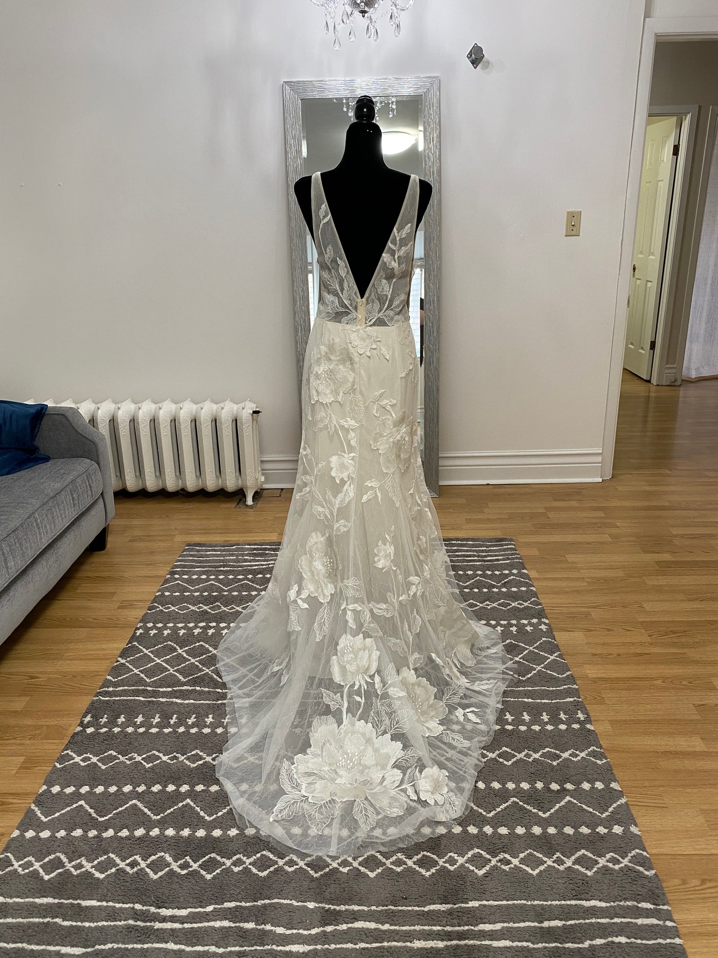 A Willowby by Watter Honor 52122 - Sample Size wedding dress with floral motifs displayed on a mannequin in a living room, reflecting in a full-length mirror.