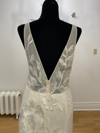 Mannequin displaying a sleeveless white wedding dress with floral motifs, featuring a deep v-neckline against a plain wall and radiator background. The dress is the Willowby by Watter Honor 52122 - Sample Size from Bergamot Bridal.
