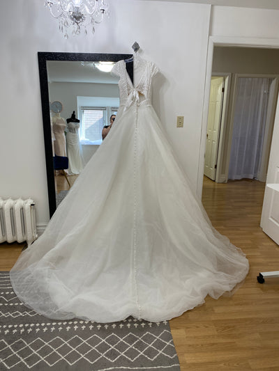 A Bergamot Bridal Allure Disney Wedding Dress - Rapunzel - Off The Rack displayed in a room with a mirror reflecting its back.