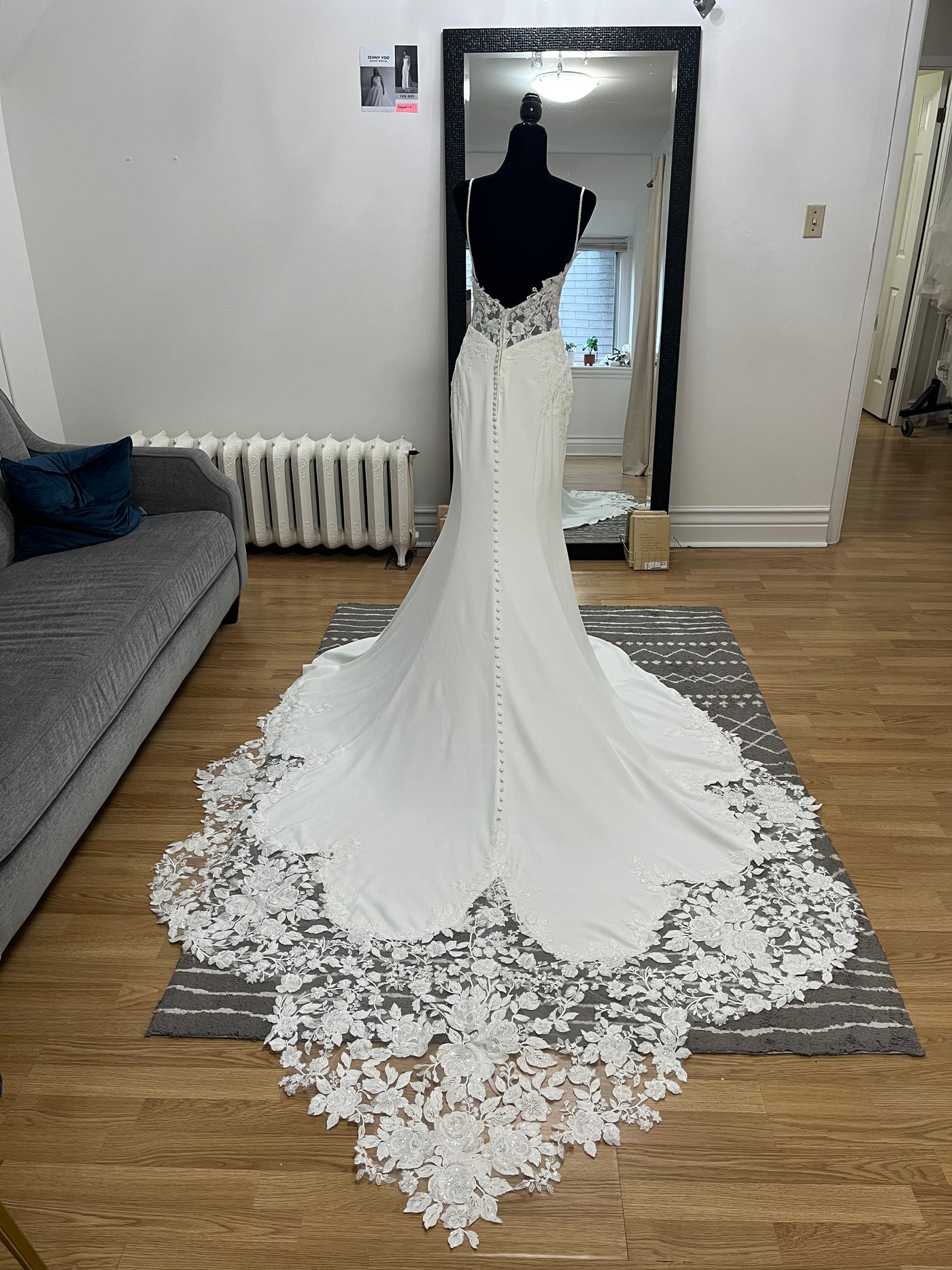 The Bergamot Bridal Crepe and Lace Fit & Flare Gown with lace train and beaded spaghetti straps is displayed on a black mannequin before a large mirror in the living room.