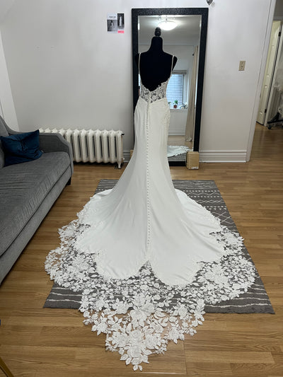 The Bergamot Bridal Crepe and Lace Fit & Flare Gown with lace train and beaded spaghetti straps is displayed on a black mannequin before a large mirror in the living room.