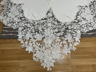 The Crepe and Lace Fit & Flare Gown with lace train and beaded spaghetti straps by Bergamot Bridal features intricate white floral lace embroidery, ideal for wedding dresses, displayed on a wooden floor over a striped gray and white rug.