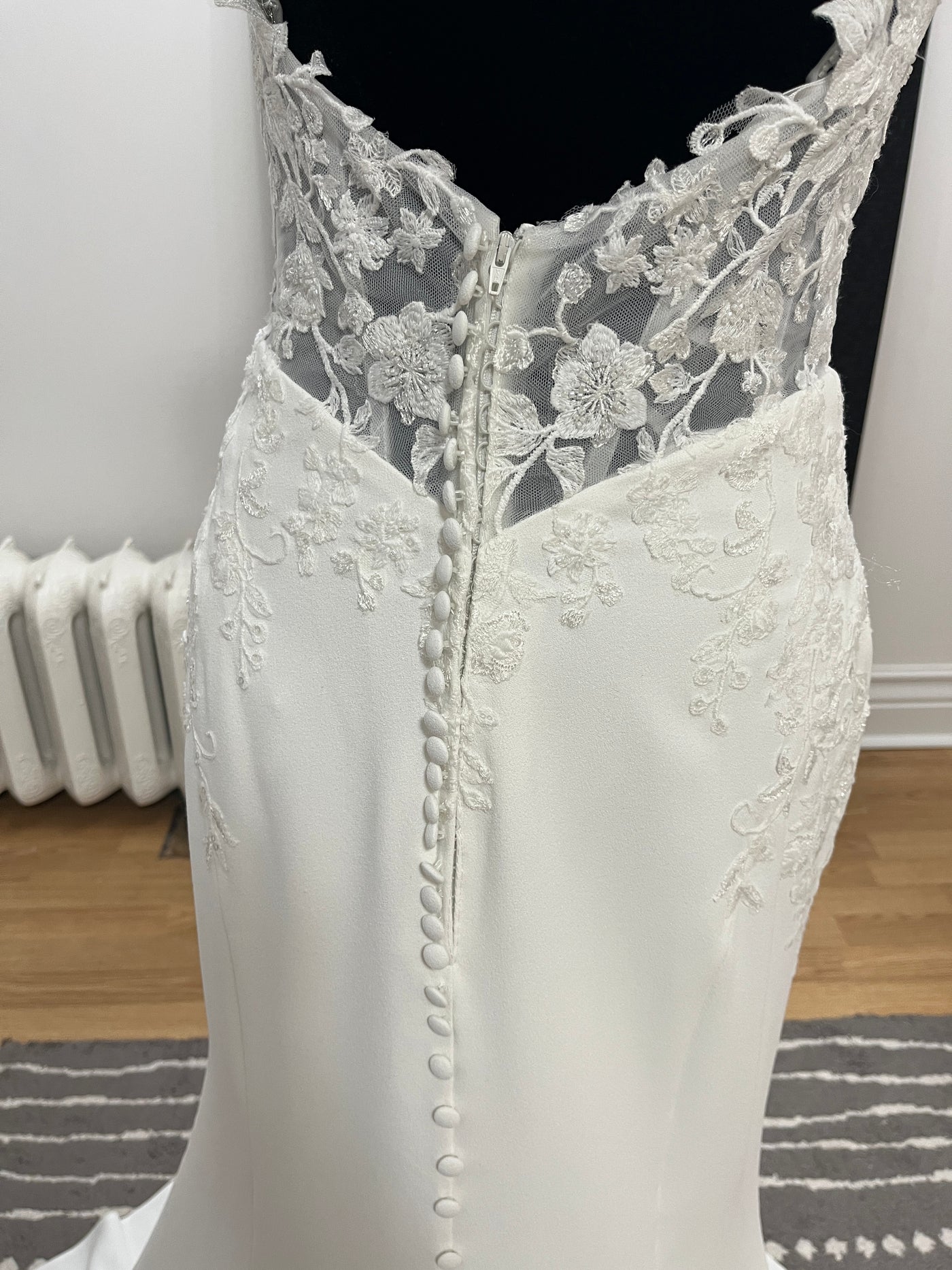 Close-up of the Bergamot Bridal Crepe and Lace Fit & Flare Gown on a mannequin, showcasing beaded spaghetti straps, fabric buttons, floral embroidery, and a scalloped lace train.