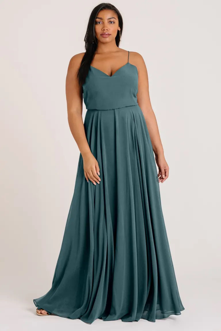 A woman in the Inesse Bridesmaid Dress by Jenny Yoo from Bergamot Bridal, featuring a floor-length teal chiffon fabric, spaghetti straps, and a circle skirt, stands against a plain background.