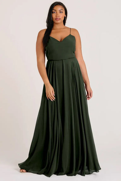 A woman is wearing the Inesse Bridesmaid Dress by Jenny Yoo from Bergamot Bridal, a floor-length, dark green chiffon dress with thin straps and a V-neck. One hand rests by her side while the other gently touches her waist. She stands against a plain background, letting the circle skirt gracefully flow around her.