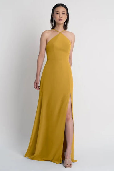 Woman posing in a sleek mustard yellow Ingrid bridesmaid dress by Jenny Yoo with a halter neckline and a side slit from Bergamot Bridal.