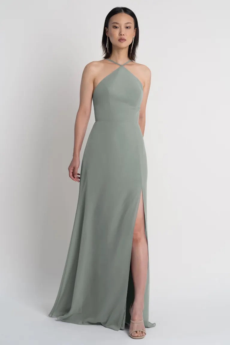 A woman modeling an elegant sage green Ingrid bridesmaid dress by Jenny Yoo with a pointed halter neckline and a side slit from Bergamot Bridal.