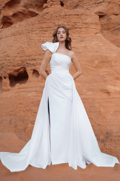 A woman in an elegant Isla - Jenny Yoo Wedding Dress gown with a dramatic shoulder detail stands against a red rock formation from Bergamot Bridal.