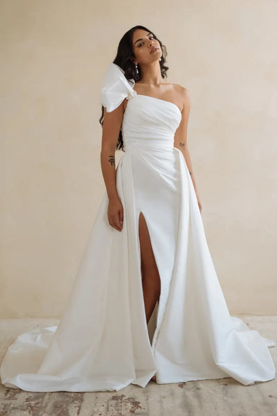 Woman in an elegant white Isla - Jenny Yoo Wedding Dress with a slit and train, posing against a neutral backdrop from Bergamot Bridal.
