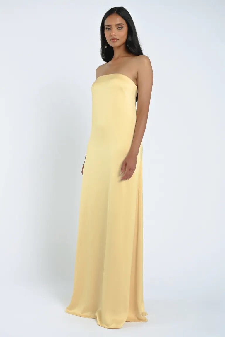 A woman stands wearing the James Bridesmaid Dress by Jenny Yoo from Bergamot Bridal, featuring a strapless neckline and crafted from luxe satin fabric, against a plain white background.