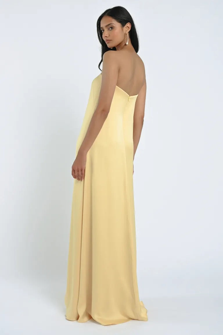 A woman with long dark hair stands sideways and looks over her shoulder, wearing a strapless, floor-length James Bridesmaid Dress by Jenny Yoo in pale yellow, made from luxe satin fabric by Bergamot Bridal.