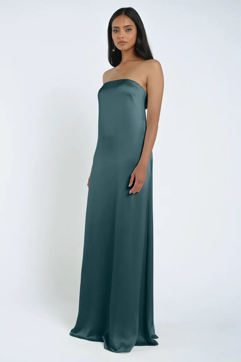 A woman stands wearing a floor-length, strapless James Bridesmaid Dress by Jenny Yoo in teal, made from smooth luxe satin fabric with a refined sheath silhouette. She is positioned against a plain white background. The elegant dress is from the brand Bergamot Bridal.