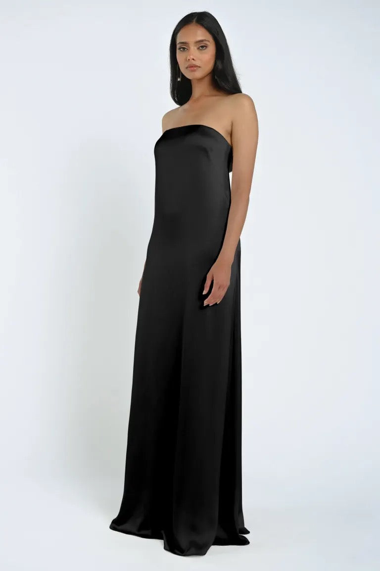 A woman stands wearing a long, strapless black James Bridesmaid Dress by Jenny Yoo, featuring luxurious satin fabric and a sleek sheath silhouette, against a plain backdrop.