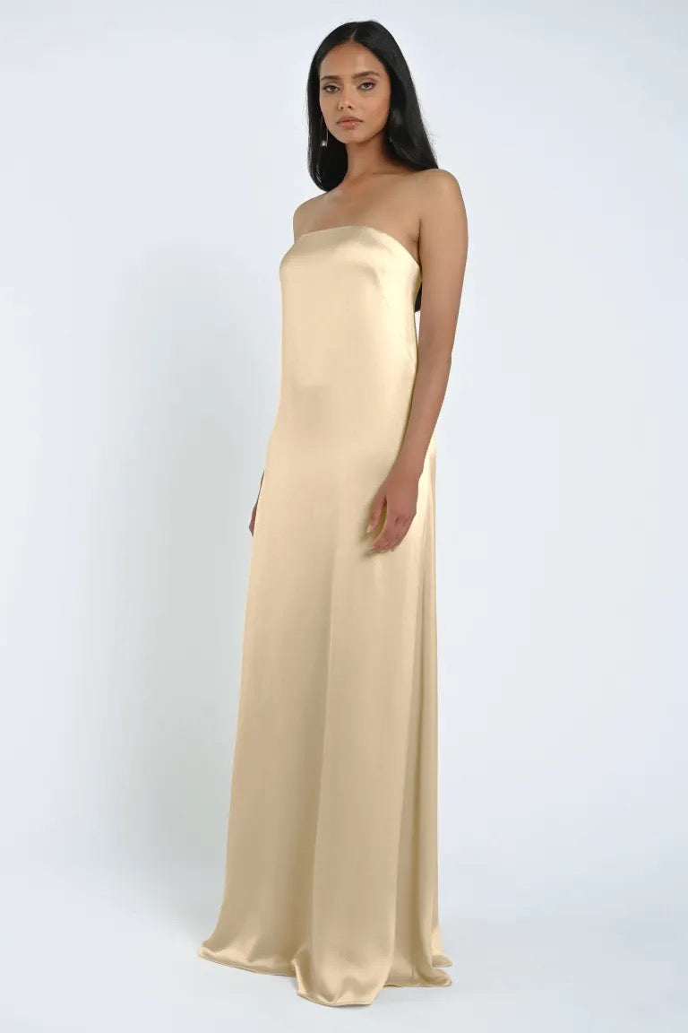 A woman in a sleeveless, strapless James Bridesmaid Dress by Jenny Yoo made of luxe satin fabric from Bergamot Bridal stands in front of a plain white background.