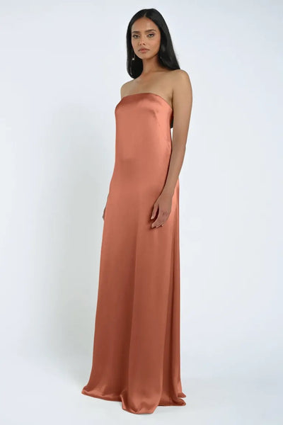 A woman in a James Bridesmaid Dress by Jenny Yoo from Bergamot Bridal, featuring a strapless neckline and floor-length luxe satin fabric in rust, stands against a plain white background.