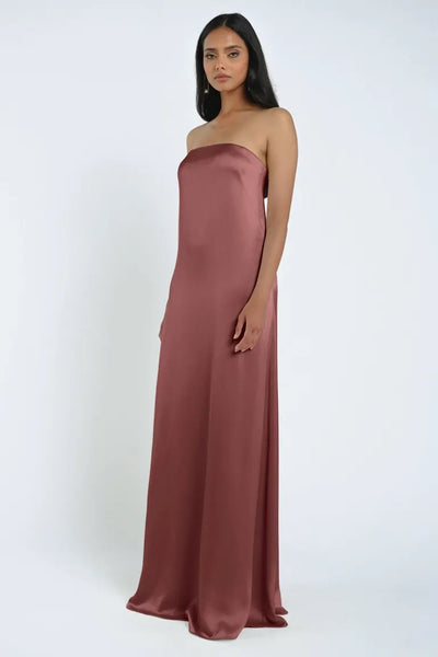 A woman stands against a plain background wearing the Bergamot Bridal James Bridesmaid Dress by Jenny Yoo, featuring a strapless neckline and flowing in floor-length, luxe satin fabric.