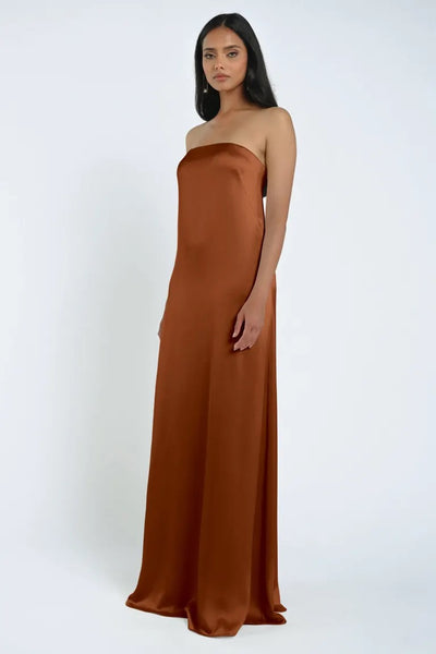 A woman stands against a plain background wearing the James Bridesmaid Dress by Jenny Yoo from Bergamot Bridal, a floor-length, strapless gown crafted from luxe satin fabric in an elegant brown hue.