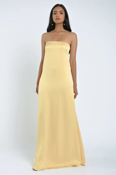 A woman stands wearing a strapless, floor-length, light yellow James Bridesmaid Dress by Jenny Yoo from Bergamot Bridal, crafted from luxe satin fabric against a plain white background.