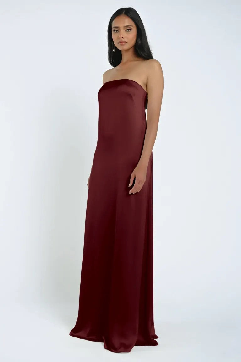 A woman stands in a strapless, floor-length dark red James Bridesmaid Dress by Jenny Yoo made of luxe satin fabric against a plain white background, featured by Bergamot Bridal.