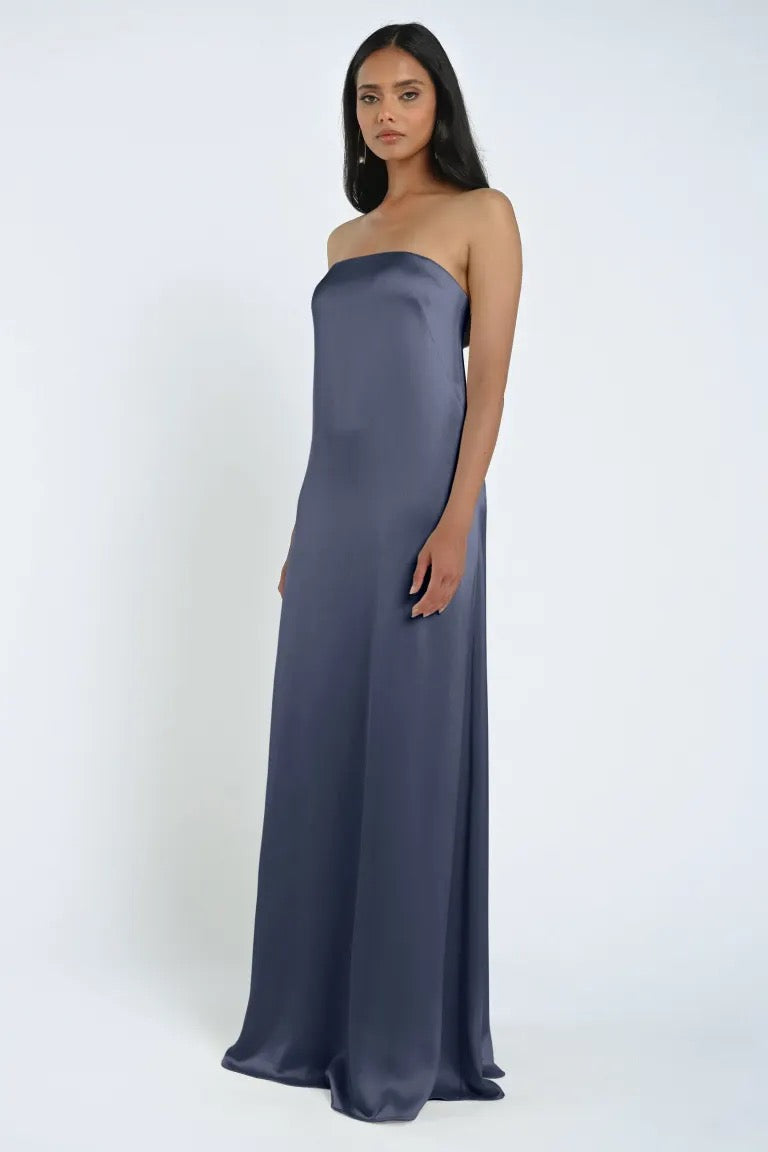 A woman stands wearing the James Bridesmaid Dress by Jenny Yoo from Bergamot Bridal, a long, strapless sheath silhouette crafted from luxe satin fabric, against a plain white background.