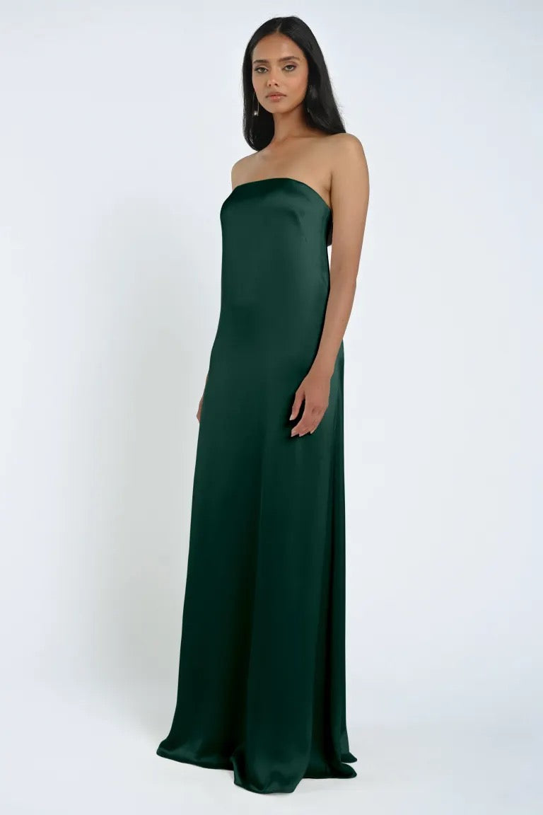 A woman stands wearing the strapless, floor-length James Bridesmaid Dress by Jenny Yoo in luxe satin fabric, its rich green hue shimmering against a plain white background.